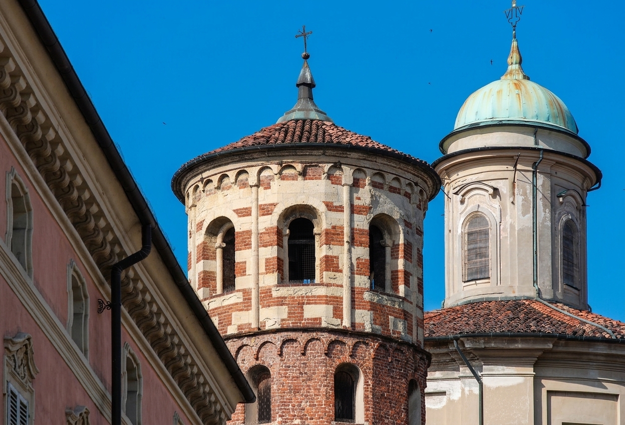 The top of Asti's Torre Rossa, banded in red brick and pale sandstone, beside the green dome of Santa Caterina against a blue sky