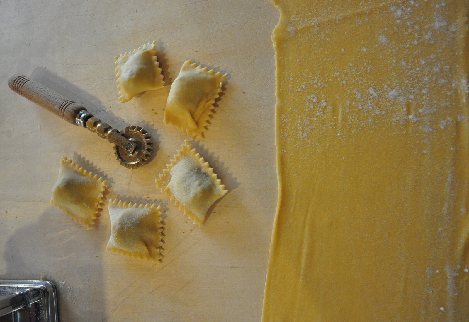 Freshly cut agnolotti and a fluted pasta wheel on a floured board beside a sheet of egg pasta