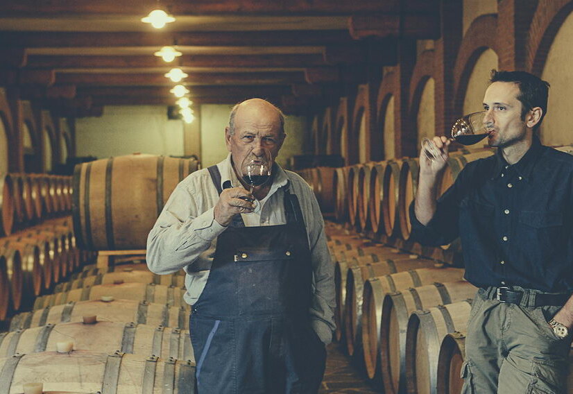 An older and a younger winemaker tasting red wine among rows of barrels in the Rovero cellar