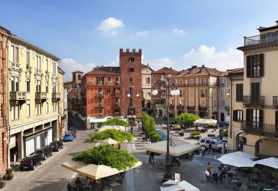 A piazza in central Asti with café umbrellas, planted trees and a medieval brick tower at its far end