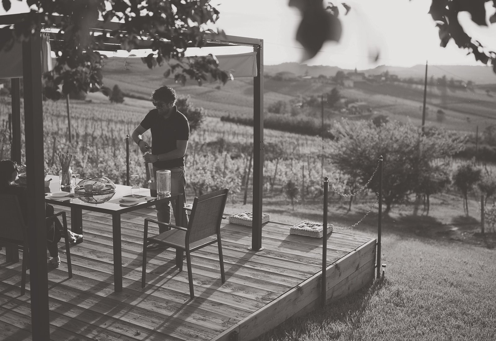 A host opening a bottle at a table on the wooden terrace above the Ca 'd Tantin vineyards in evening light (black and white)