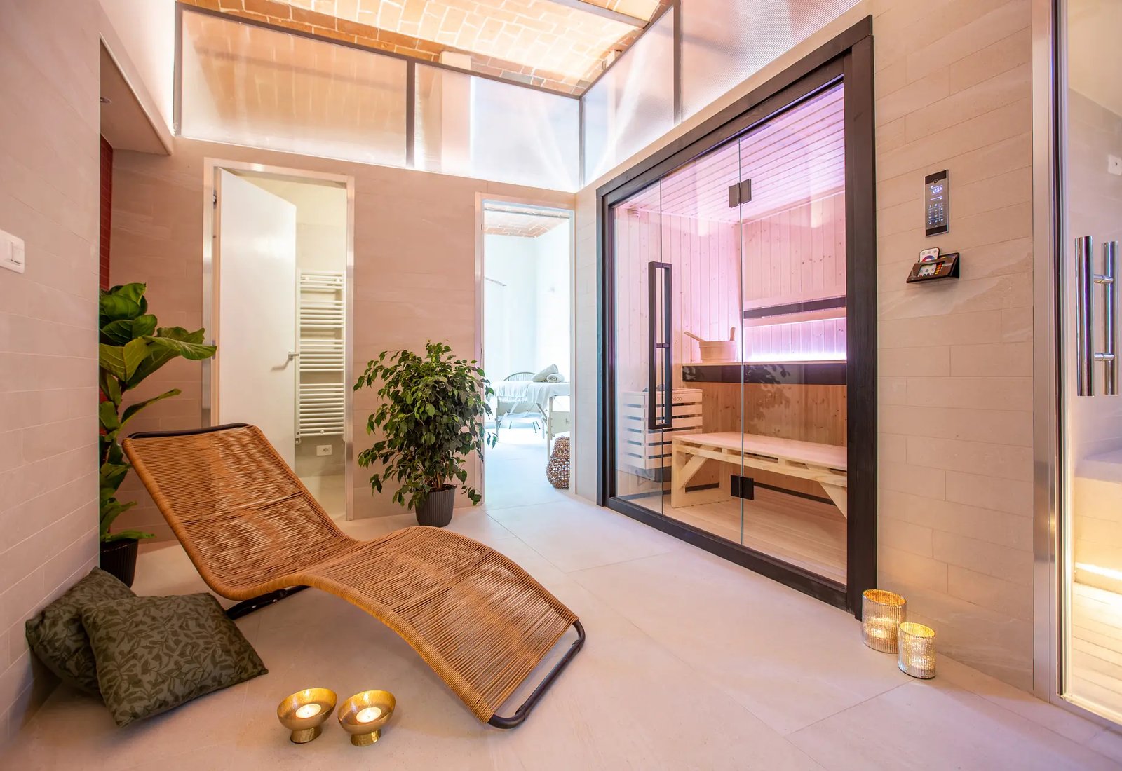 The spa's relaxation room at Retreat Monferrato: a woven lounger, candles and plants beside the glass-fronted sauna, with the massage room through the doorway
